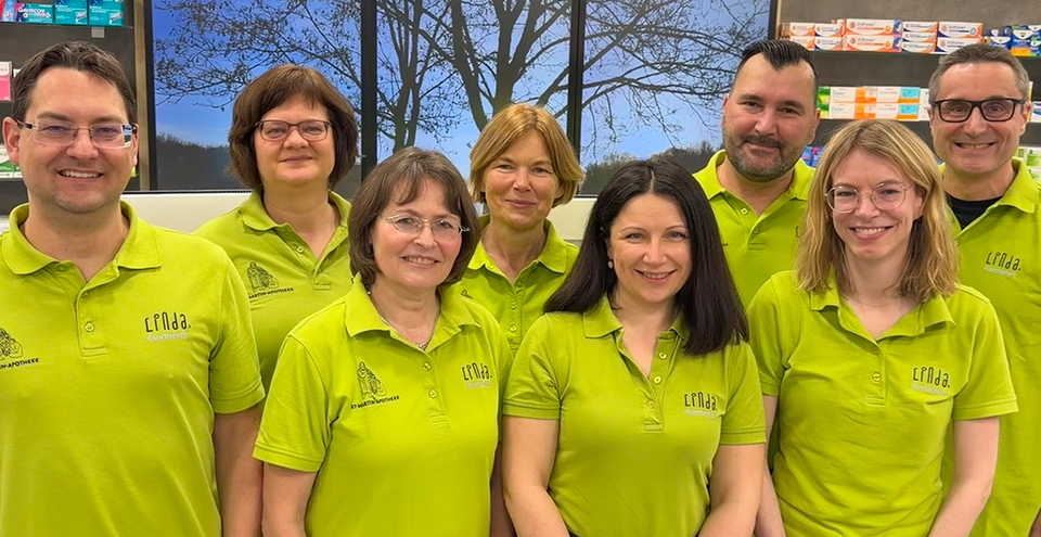 Team der St. Martin Apotheke in grünen Polo-Shirts, lächelnd in der Offizin stehend.
